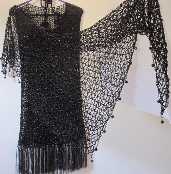 Black lurex crochet shawl draped over a black fringed dress on a hanger, side view of the shimmering wrap.
