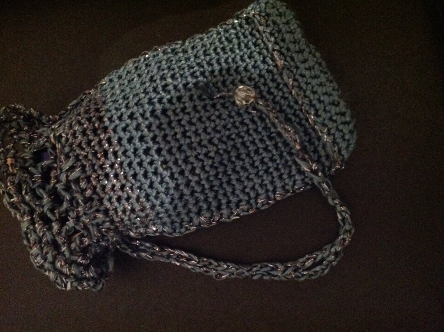 Grey crochet clutch on a dark surface, side view showing the woven strap and button detail.
