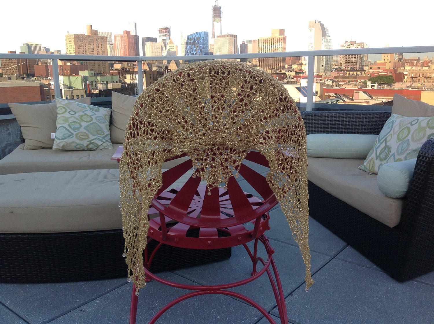 Gold shawl draped over a red metal chair on a rooftop terrace, NYC skyline and outdoor lounge seating in the background.