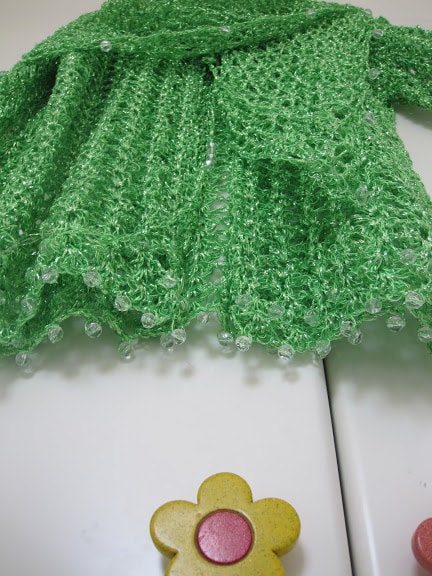 Green shawl folded on a white surface beside a flower-shaped knob, close-up of the beaded hem.