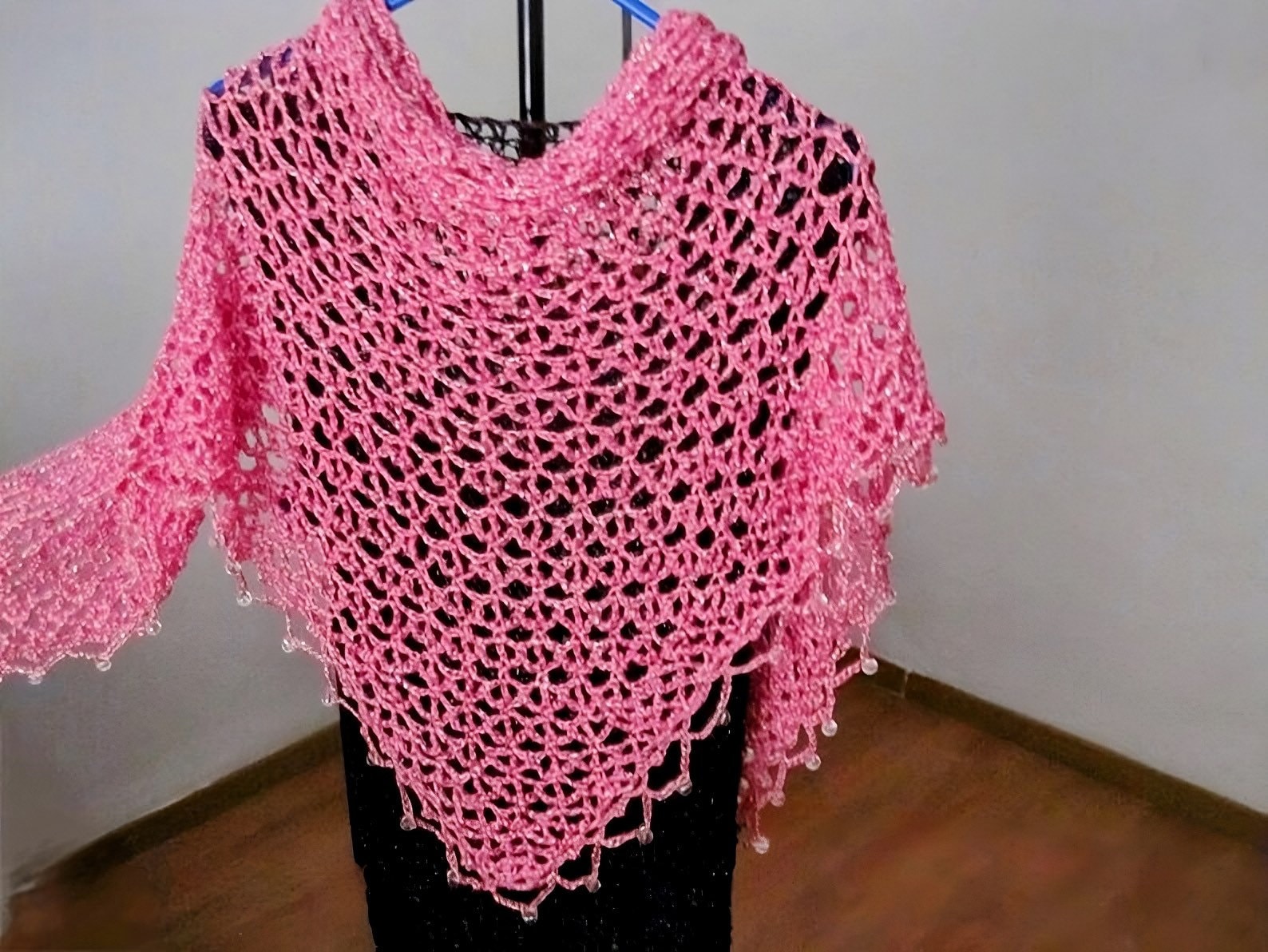 Pink lurex crochet shawl on a hanger over a black dress, full view of the shimmering openwork wrap.