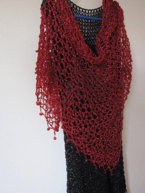 Red lurex crochet shawl draped over a black dress on a hanger, full view of the shimmering openwork wrap.