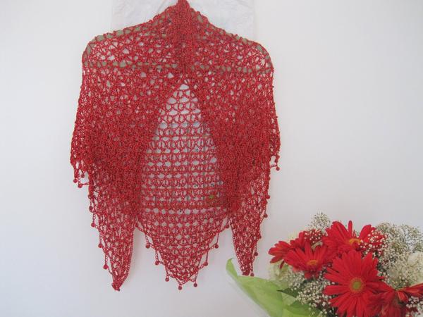 Red crochet shawl displayed flat against a white wall beside a bouquet of red flowers, showing its full triangular shape.