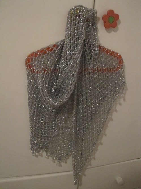 Silver shawl on an orange hanger, front view of the openwork crochet and shimmering weave.