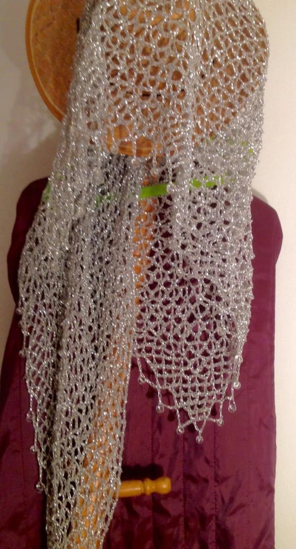 Silver lurex crochet shawl hanging on a wooden stand beneath a straw hat, long shimmering drape over a burgundy garment.