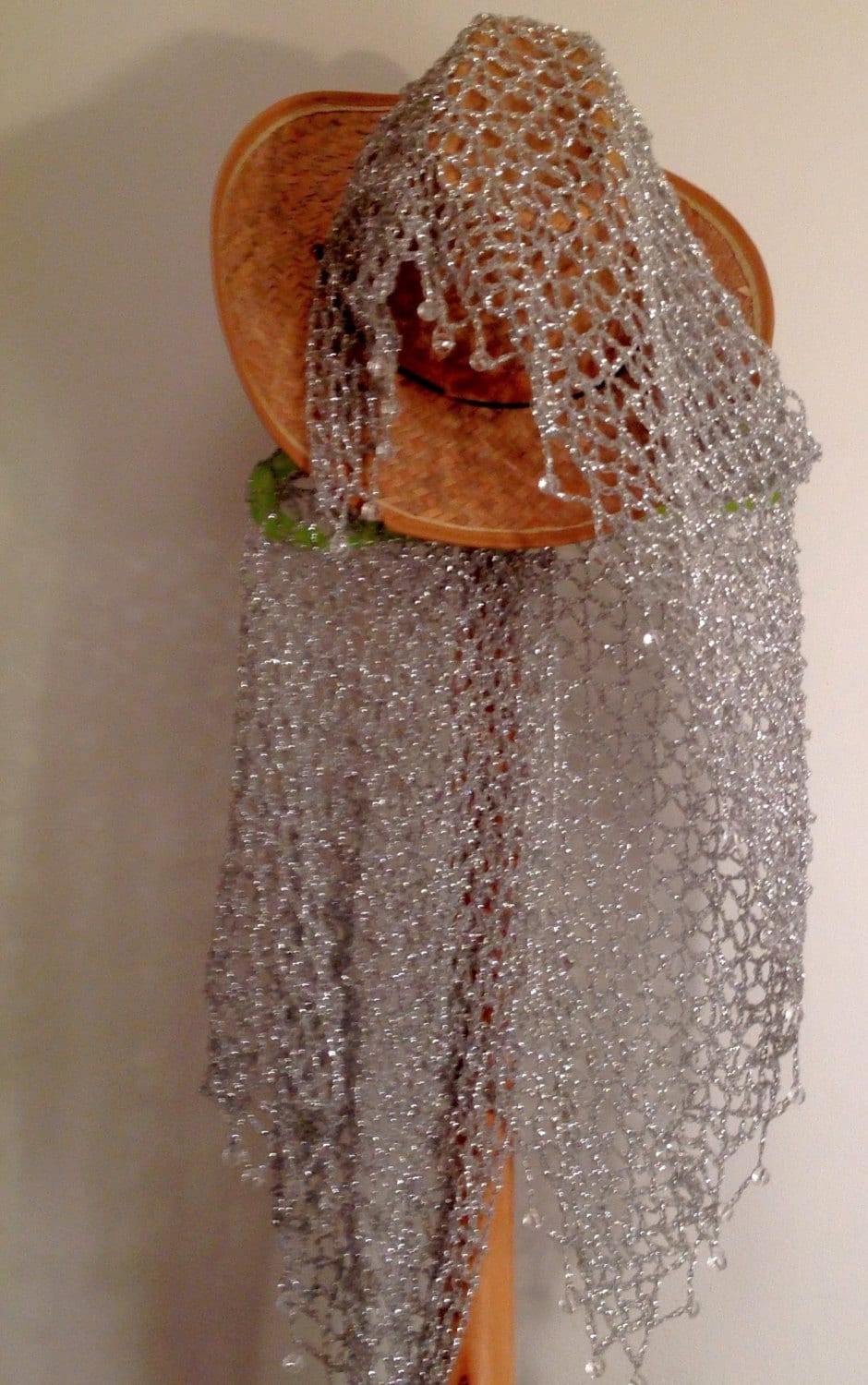 Silver lurex shawl folded over a straw hat on the stand, showing the airy crochet pattern and delicate bead trim.