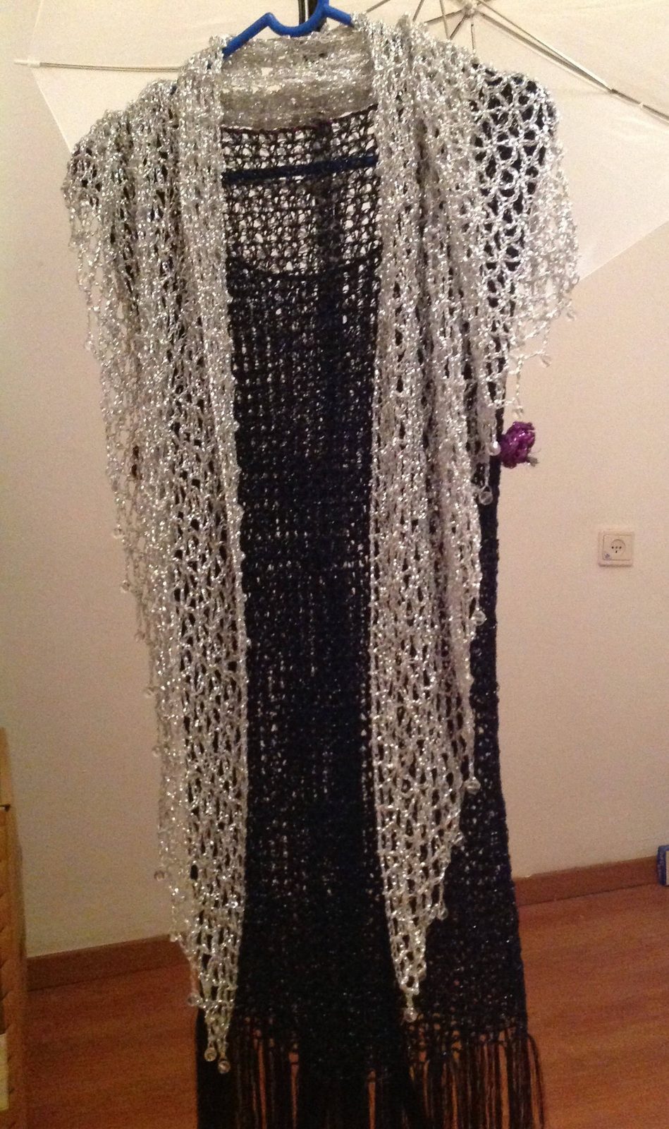 Silver lurex crochet shawl layered over a black fringed dress on a hanger, full-length view of the sparkling wrap.