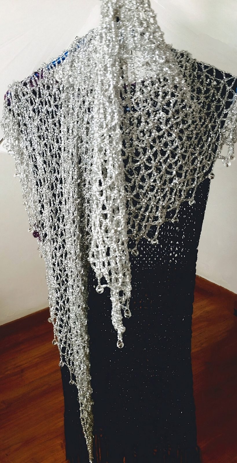 Silver beaded shawl draped over a black dress on a hanger, side view of its long flowing drape.