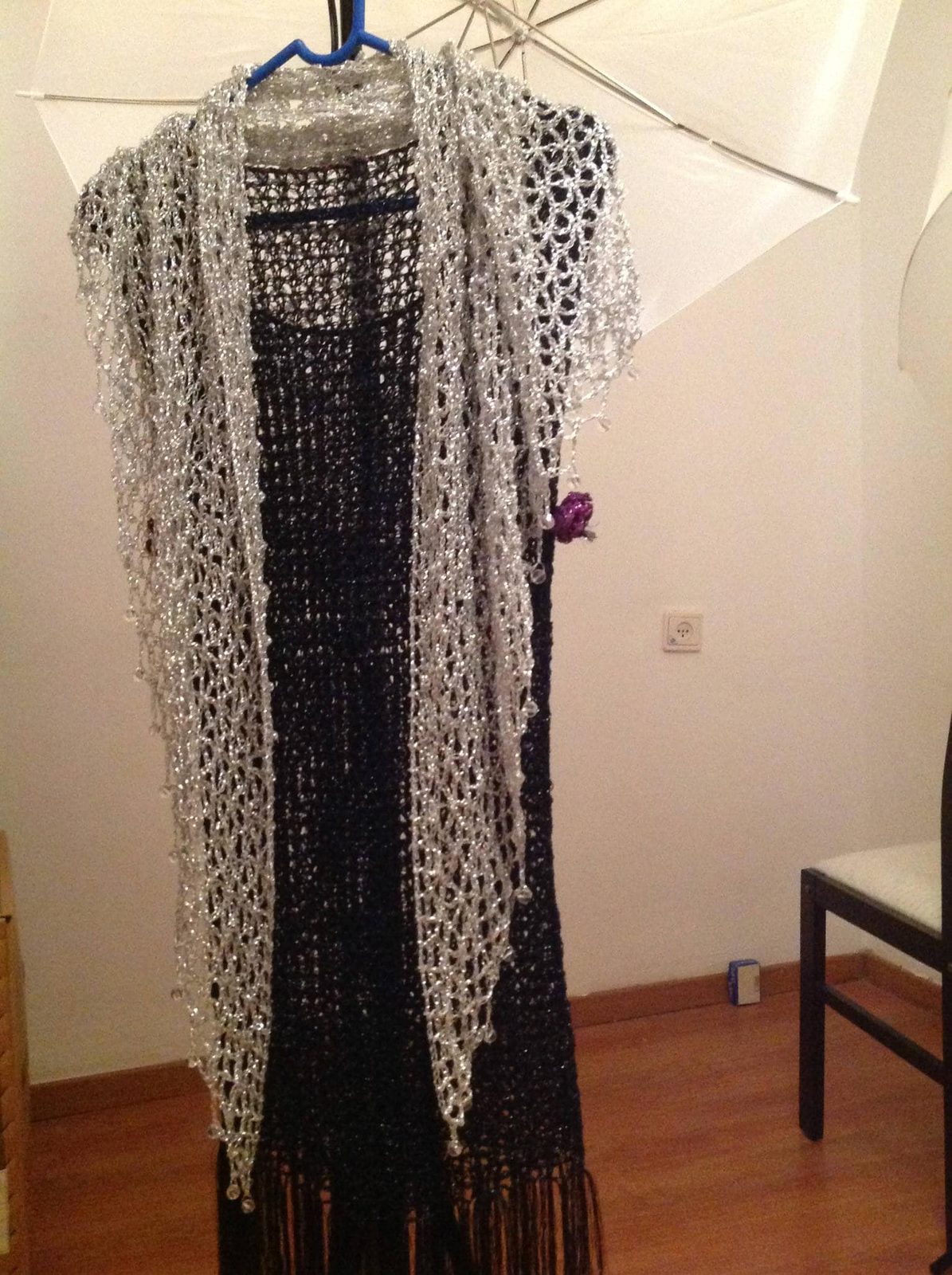 Silver shawl layered over a black fringed dress on a hanger, front view of the sparkling openwork wrap.