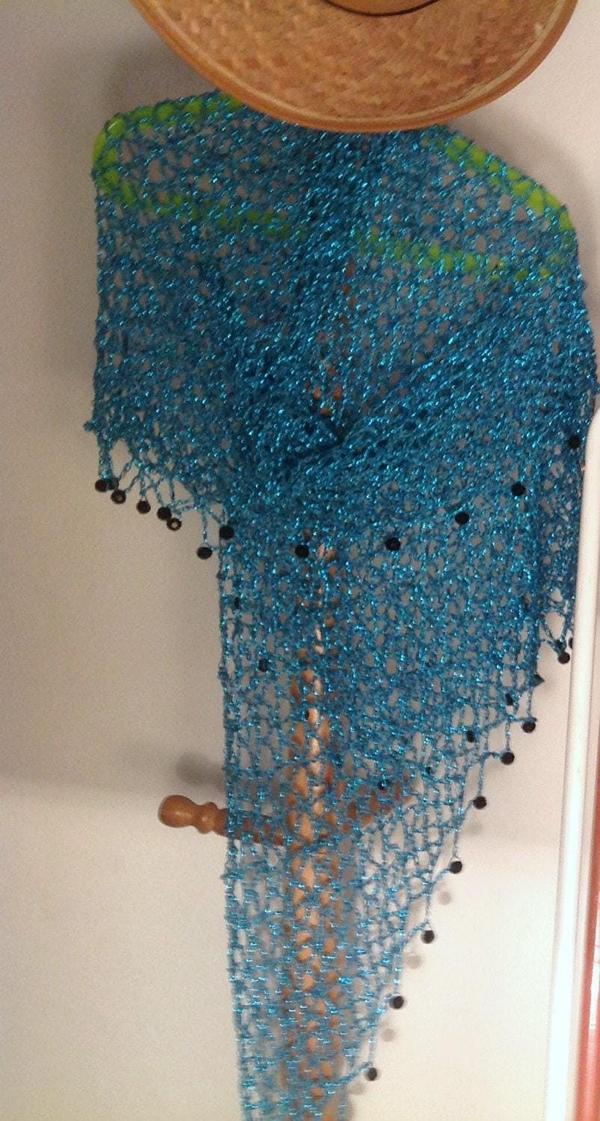 Turquoise lurex crochet shawl hanging beneath a straw hat on a wooden stand, its long shimmering drape edged with dark beads.