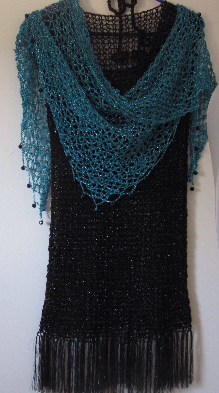 Turquoise shawl draped over a black fringed dress on a hanger, front view of the openwork wrap.